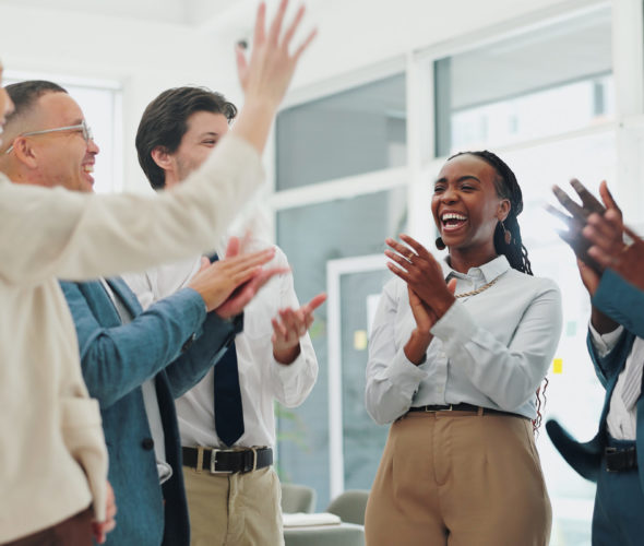 A diverse group of colleagues enthusiastically celebrates in an office, clapping and raising their hands. The scene conveys a sense of shared success and positive teamwork. The atmosphere is energetic and joyful.