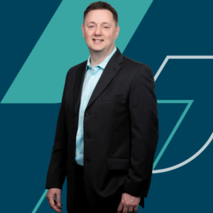 John with short hair, wearing a dark suit and a light blue shirt. He stands with a relaxed posture, smiling at the camera. The background features a modern geometric design in teal and dark blue tones.