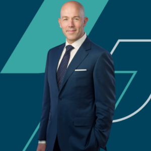 Scott is wearing a dark textured blazer, white shirt, and a dark blue tie. He has one hand in his pocket and is smiling at the camera confidently. The background features a modern geometric design in teal and dark blue tones.