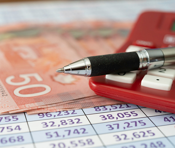 This close-up image shows a black and silver pen resting on a red calculator, which is sitting on top of a financial spreadsheet or ledger with numerical data arranged in columns. In the background, there appears to be Canadian currency, specifically what looks like a 50 dollar bill (recognizable by its reddish-pink color). The scene depicts tools typically used for financial analysis, accounting, or budgeting work - capturing the essence of financial management and data analysis.